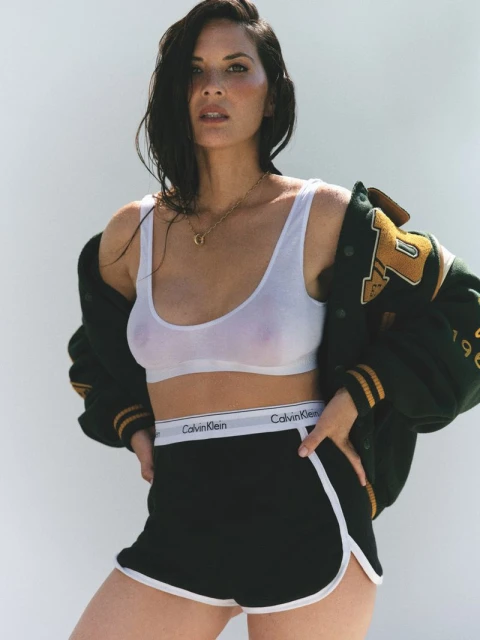 Portrait of Olivia Munn posing in a white Calvin Klein top and black shorts with a green letterman jacket over her shoulders.