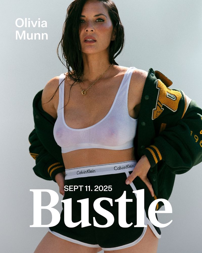 Olivia Munn poses in a sporty casual outfit consisting of a white tank top, Calvin Klein underwear, and black athletic shorts, indoor.