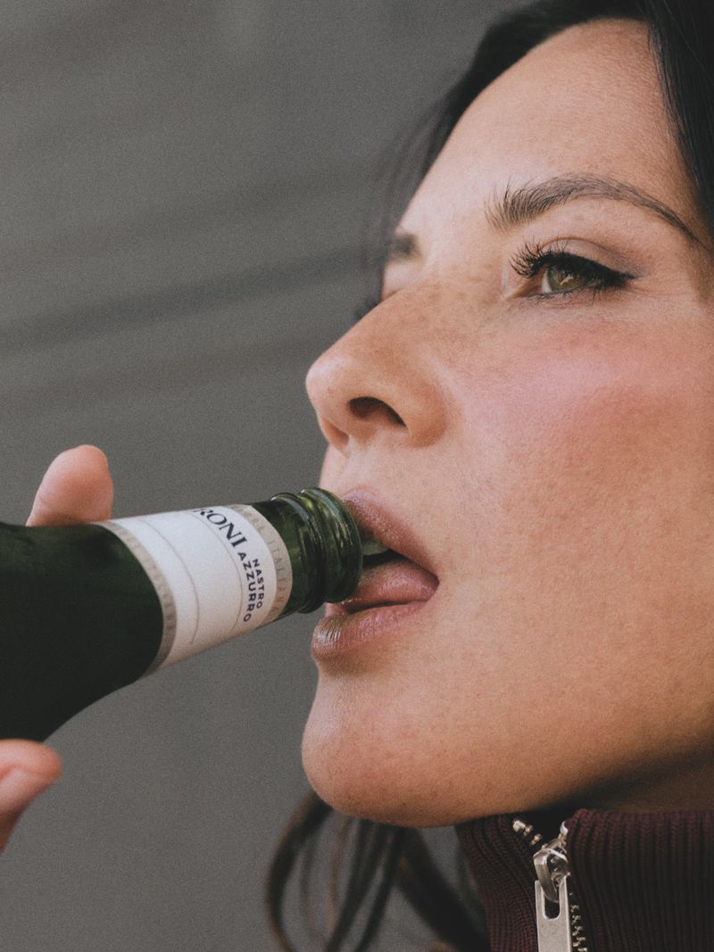 Close-up of a woman drinking directly from a green glass bottle with a white label against a grey backdrop in a high-angle shot.