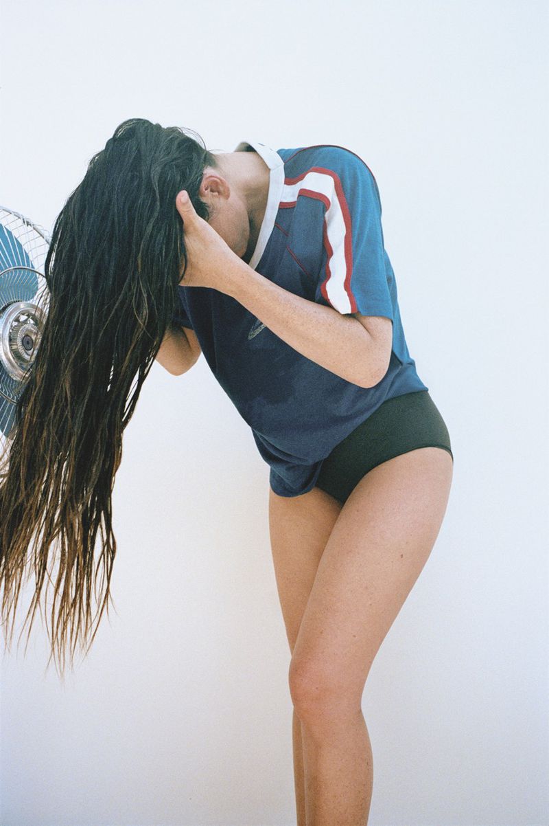Adult female with wet, long dark hair stands facing a fan wearing a blue athletic shirt against a white background in a studio shot.