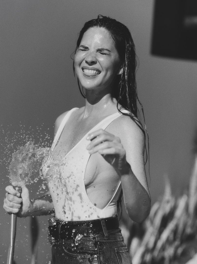 Woman in a swimsuit is smiling and getting sprayed with water from a garden hose, outdoors, with wet hair and water droplets visible.