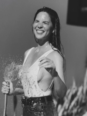 Woman in a swimsuit is smiling and getting sprayed with water from a garden hose, outdoors, with wet hair and water droplets visible.