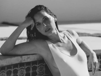 Olivia Munn posing outdoors in a light-colored swimsuit with wet hair against an architectural pool edge.