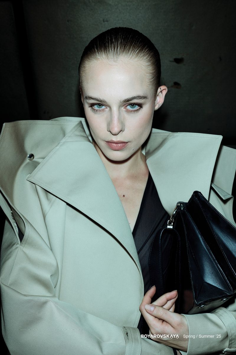 Model in structured light-colored coat holding a black handbag against a dark background for Boyarovskaya SS25 collection.