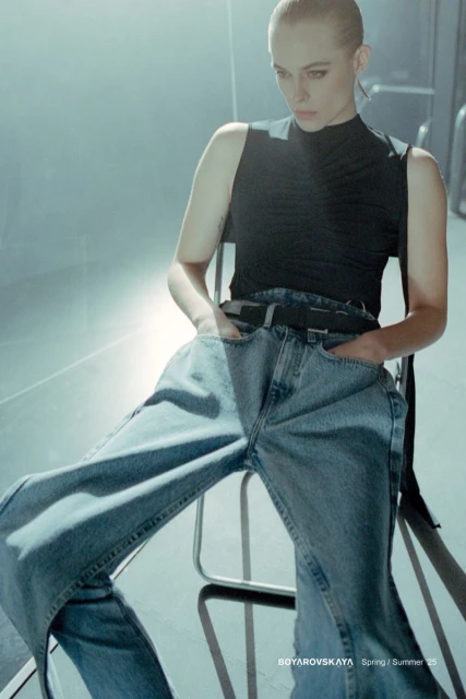 Fashion shot of a woman in a black top and denim pants sitting on a metal chair in an illuminated photo studio.