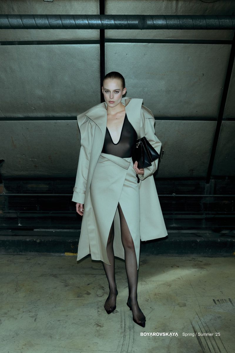 Model in cream coat and sheer black bodysuit holding a black handbag in an underground industrial space.