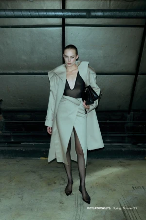 Model in cream coat and sheer black bodysuit holding a black handbag in an underground industrial space.