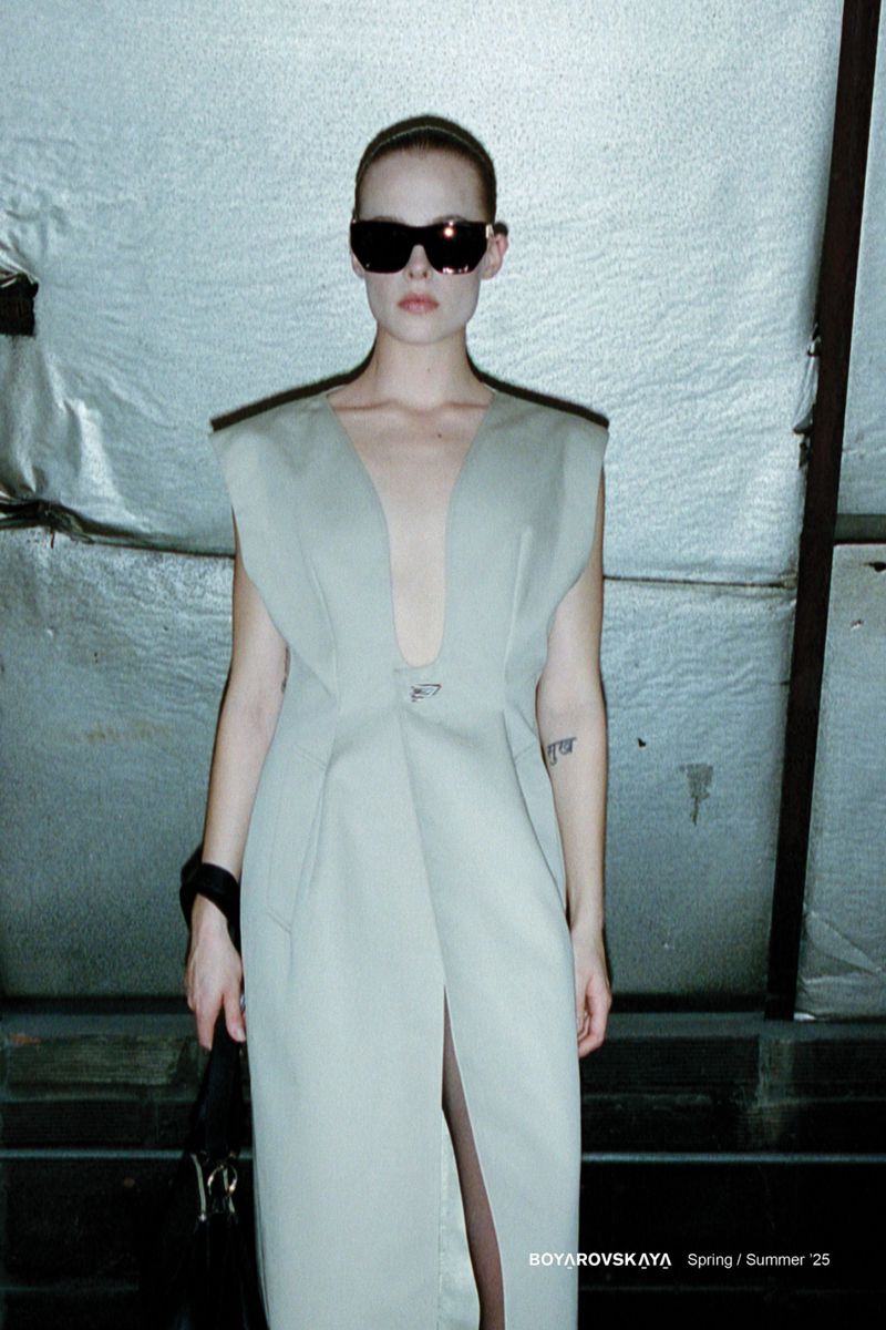 Fashion-forward woman in a light-grey maxi dress and sunglasses stands against a textured metallic backdrop, accessorized with a black handbag.