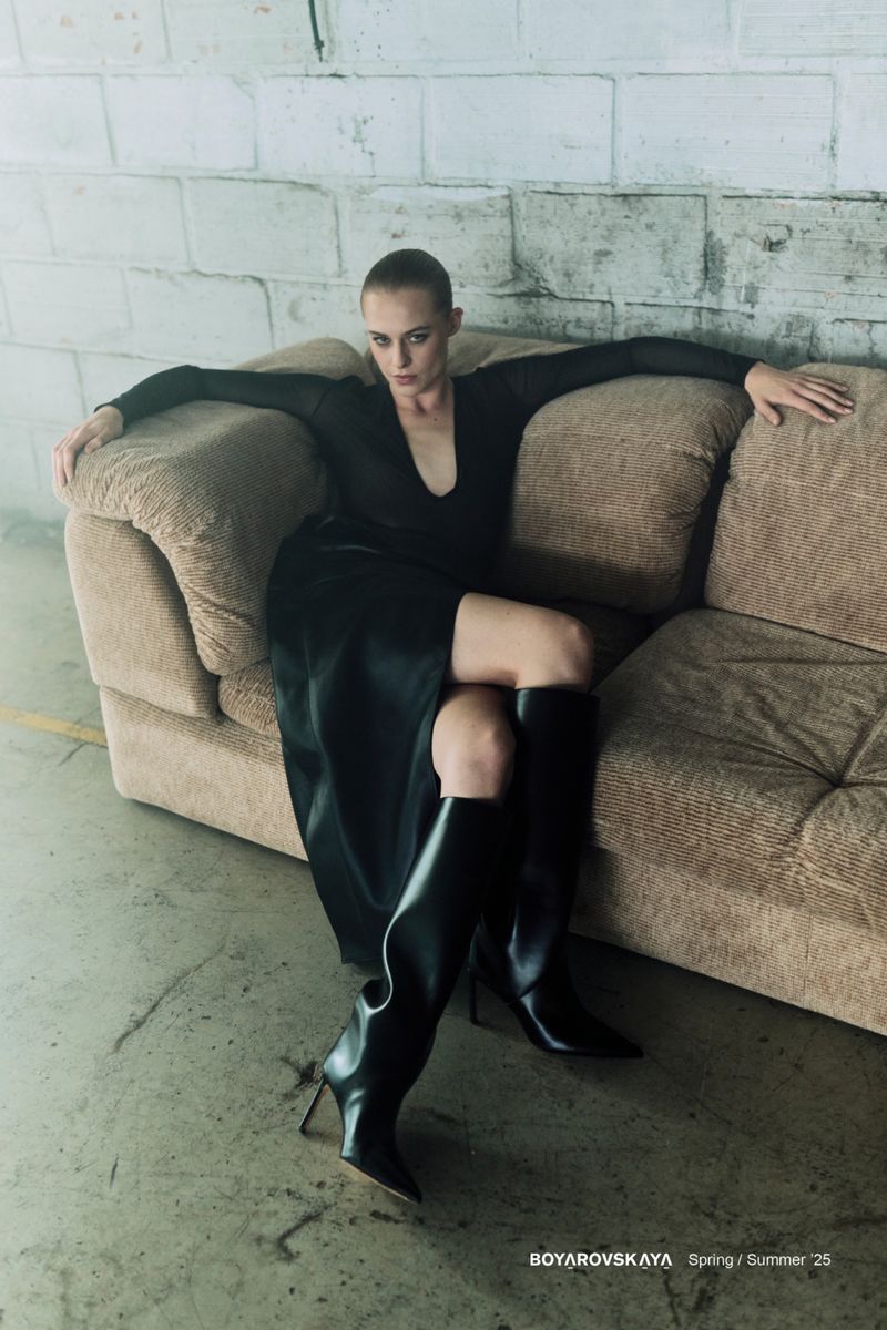 A woman in a black dress and tall black boots poses on a couch in an industrial setting with a brick wall, moody lighting.