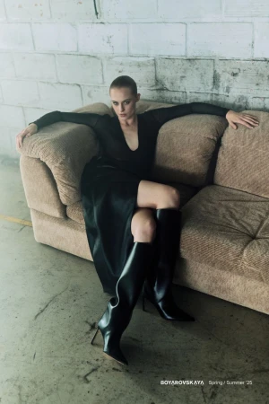 A woman in a black dress and tall black boots poses on a couch in an industrial setting with a brick wall, moody lighting.