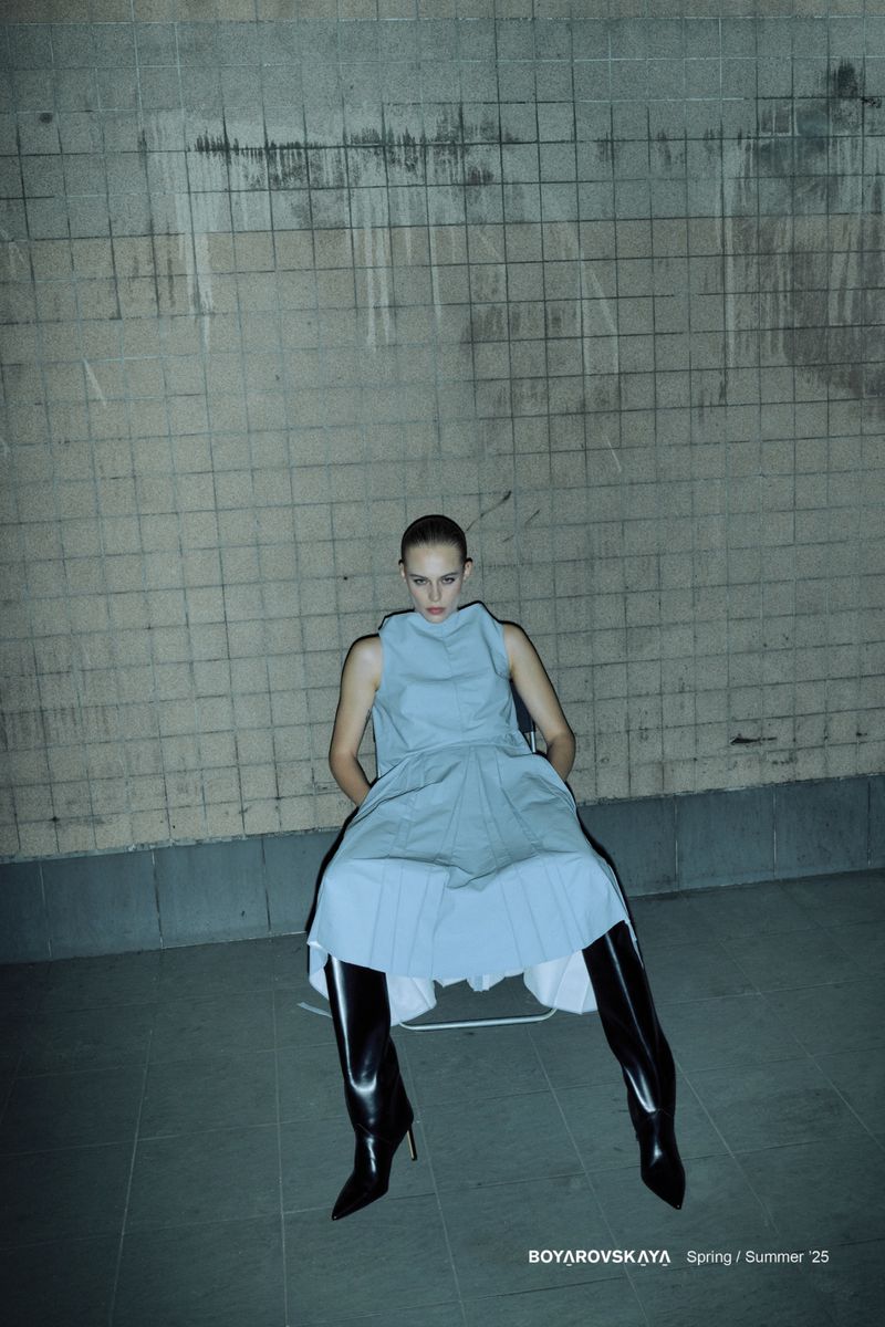 Fashion shot of a woman in a light blue dress and black patent leather boots sitting in front of a concrete block wall.
