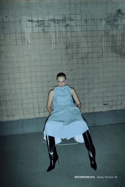 Fashion shot of a woman in a light blue dress and black patent leather boots sitting in front of a concrete block wall.