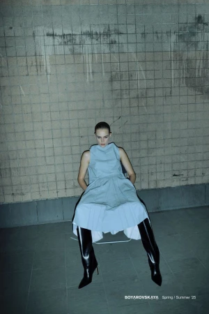 Fashion shot of a woman in a light blue dress and black patent leather boots sitting in front of a concrete block wall.