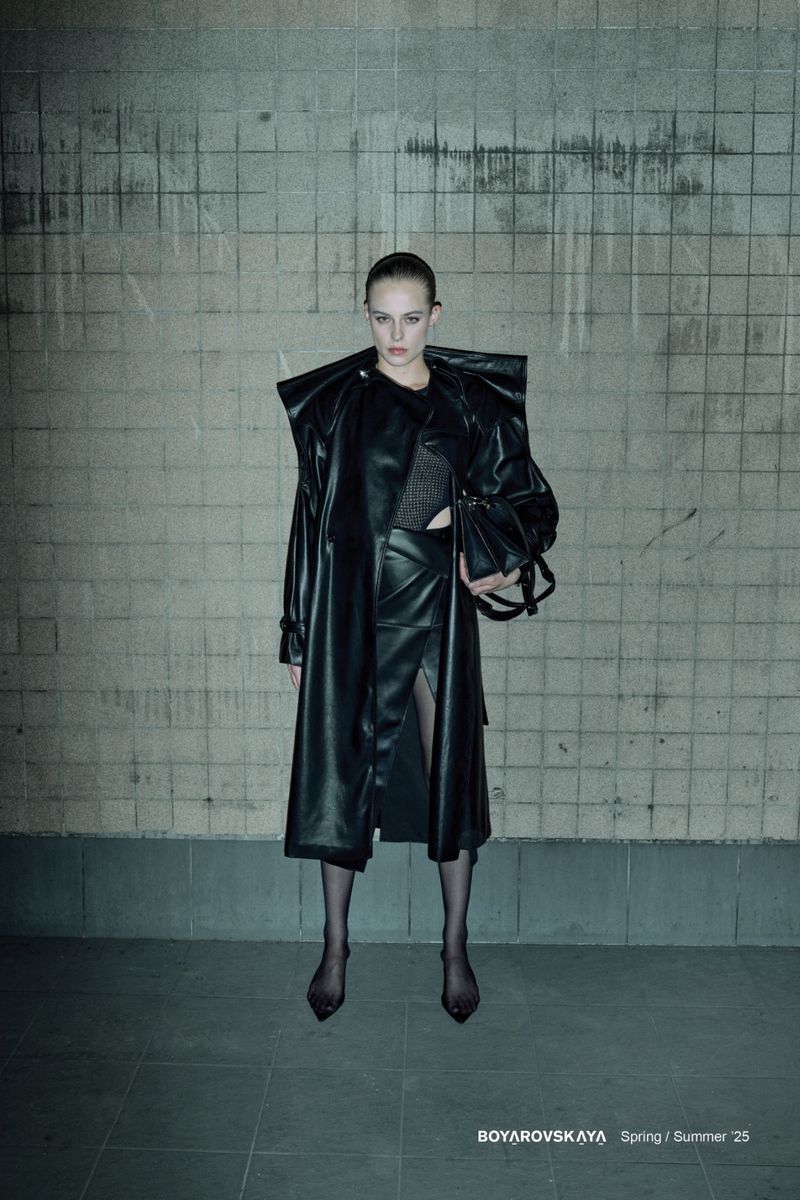 Fashion editorial image featuring a female model in a long black leather coat and skirt, posing against a textured wall in an indoor setting.