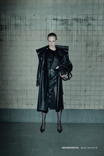 Fashion editorial image featuring a female model in a long black leather coat and skirt, posing against a textured wall in an indoor setting.