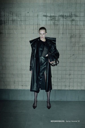 Fashion editorial image featuring a female model in a long black leather coat and skirt, posing against a textured wall in an indoor setting.