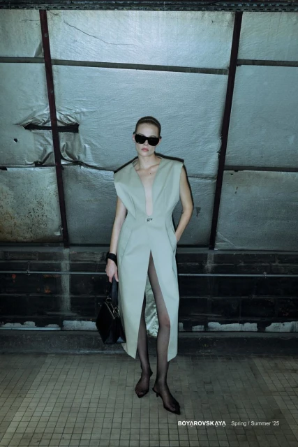 Fashion editorial shot: A woman in a sleeveless dress and sunglasses poses in front of a gridded wall, holding a black purse.