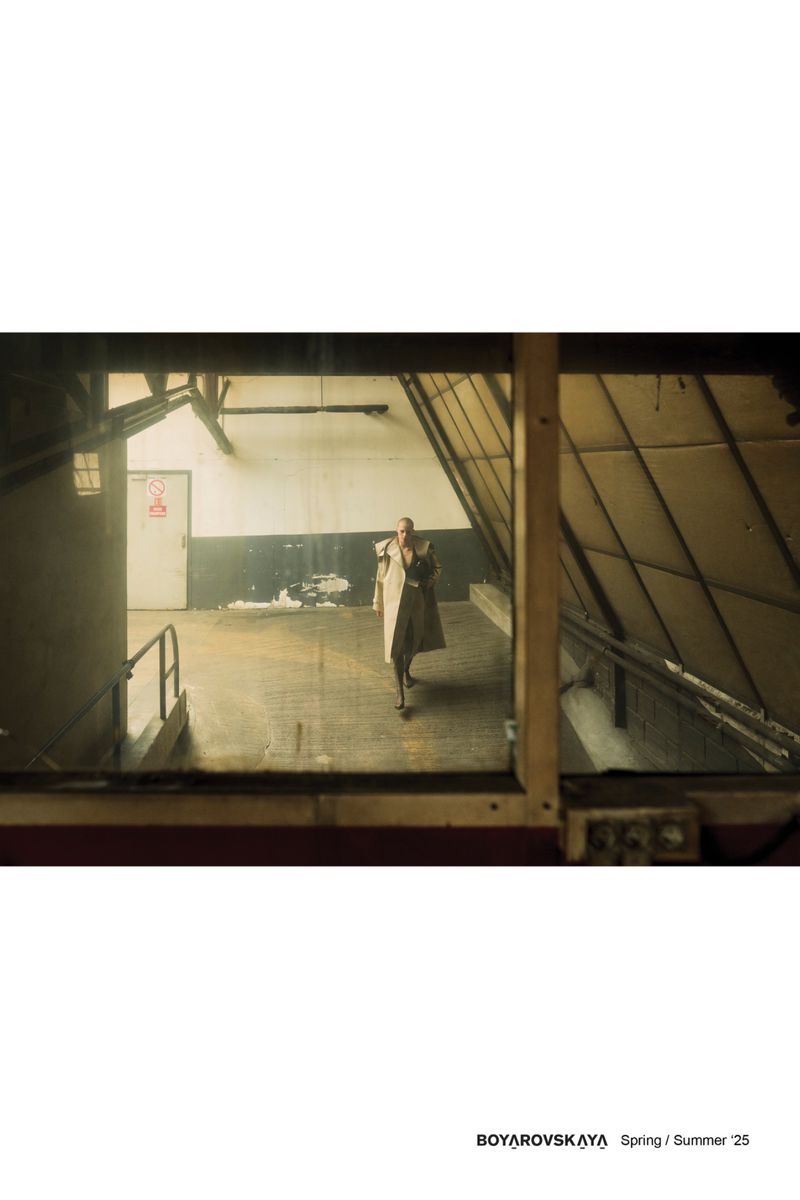 Fashion shot of a slender adult woman in a tan trench coat standing in an urban, industrial environment, taken indoors through a window.