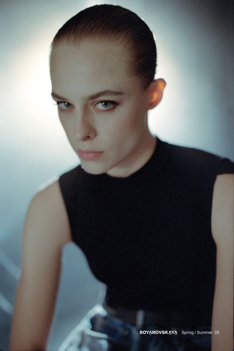 Woman with slicked-back hair poses in a sleeveless black top and denim skirt with a studio background in a medium close-up shot.