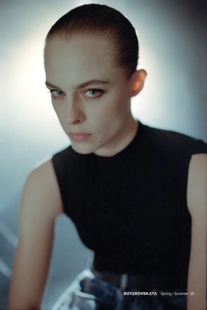 Woman with slicked-back hair poses in a sleeveless black top and denim skirt with a studio background in a medium close-up shot.