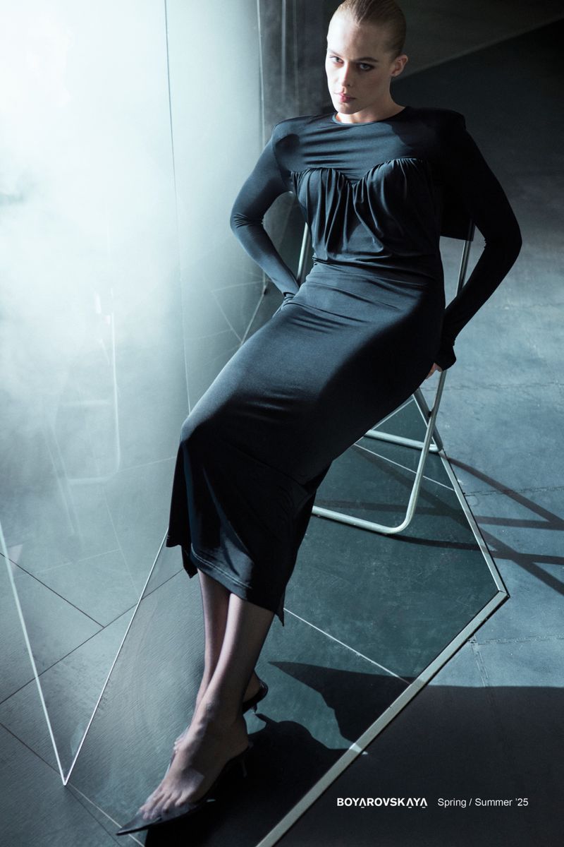 Fashion model poses in a black dress and sheer hose, sitting on a chair in a studio with geometric mirror reflections and cool lighting.