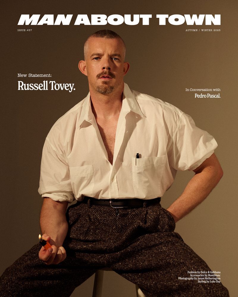 Magazine cover featuring Russell Tovey in open white shirt, dark pants, and seating on stool, holding a lighter in hand, studio shot.
