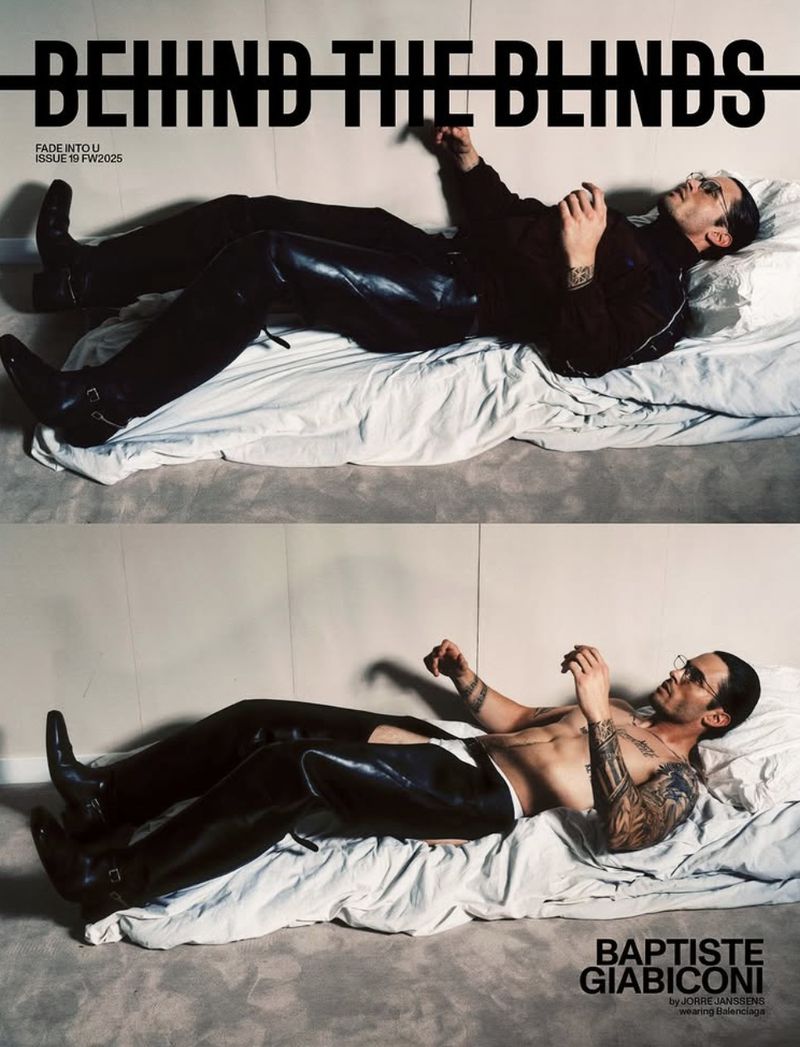 A man with tattoos is lying on a bed, wearing black clothing and black boots, in a studio shot for a magazine cover.