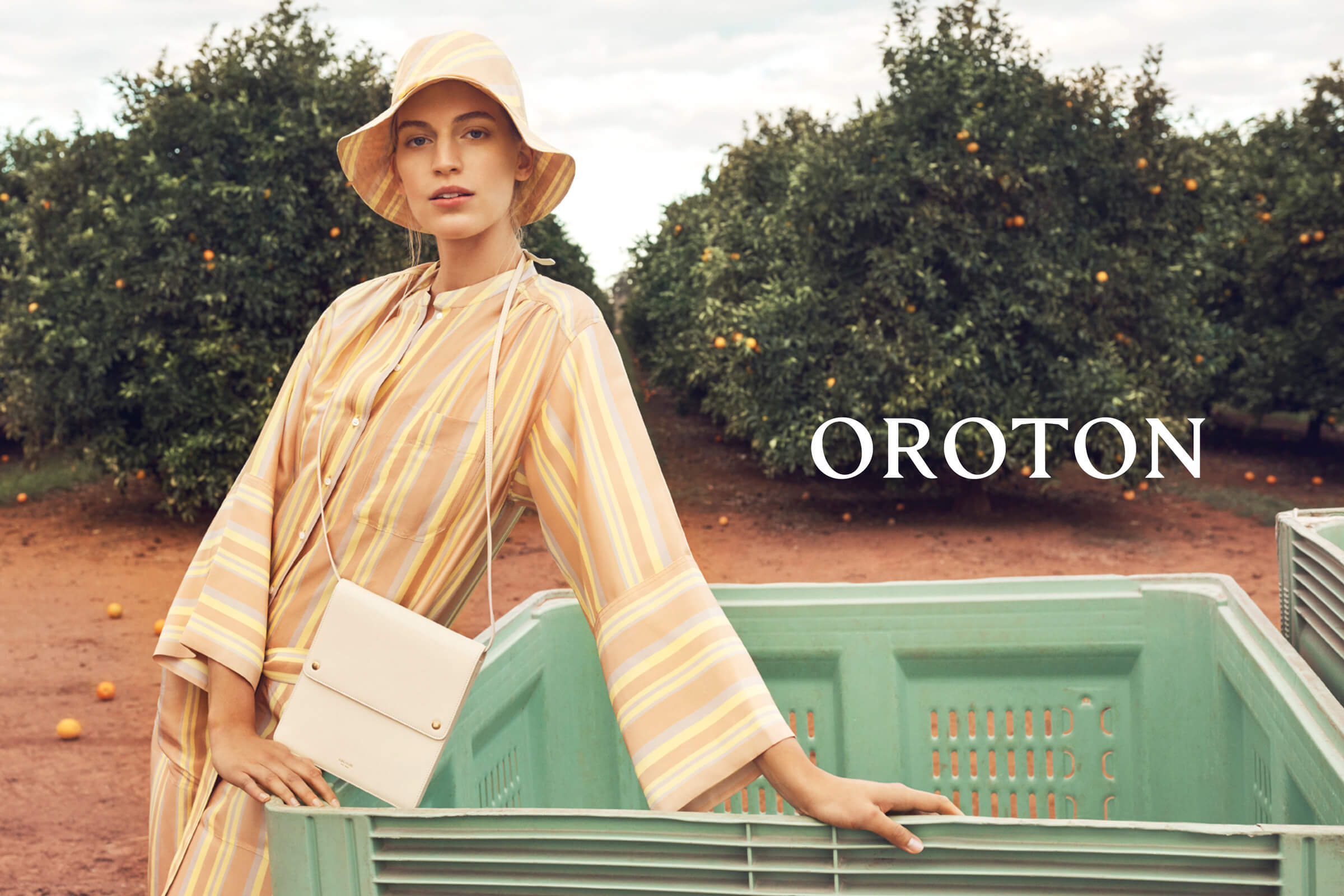 A woman in a striped dress and matching hat poses near a crate in an orange grove, holding a small crossbody bag; fashion shoot.