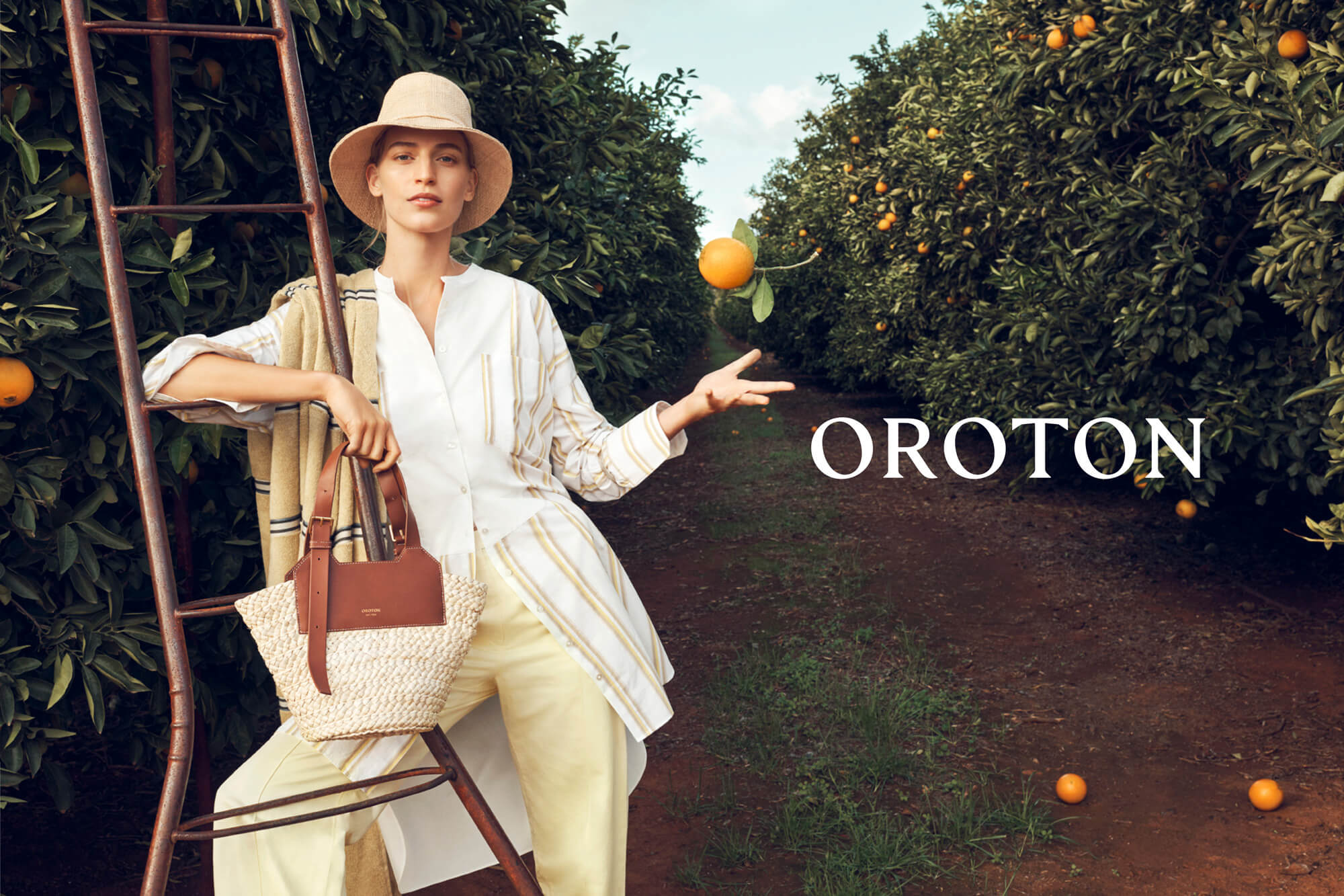 A woman in a hat stands near a ladder and orange trees, holding a purse, with the brand name Oroton in bold white letters.