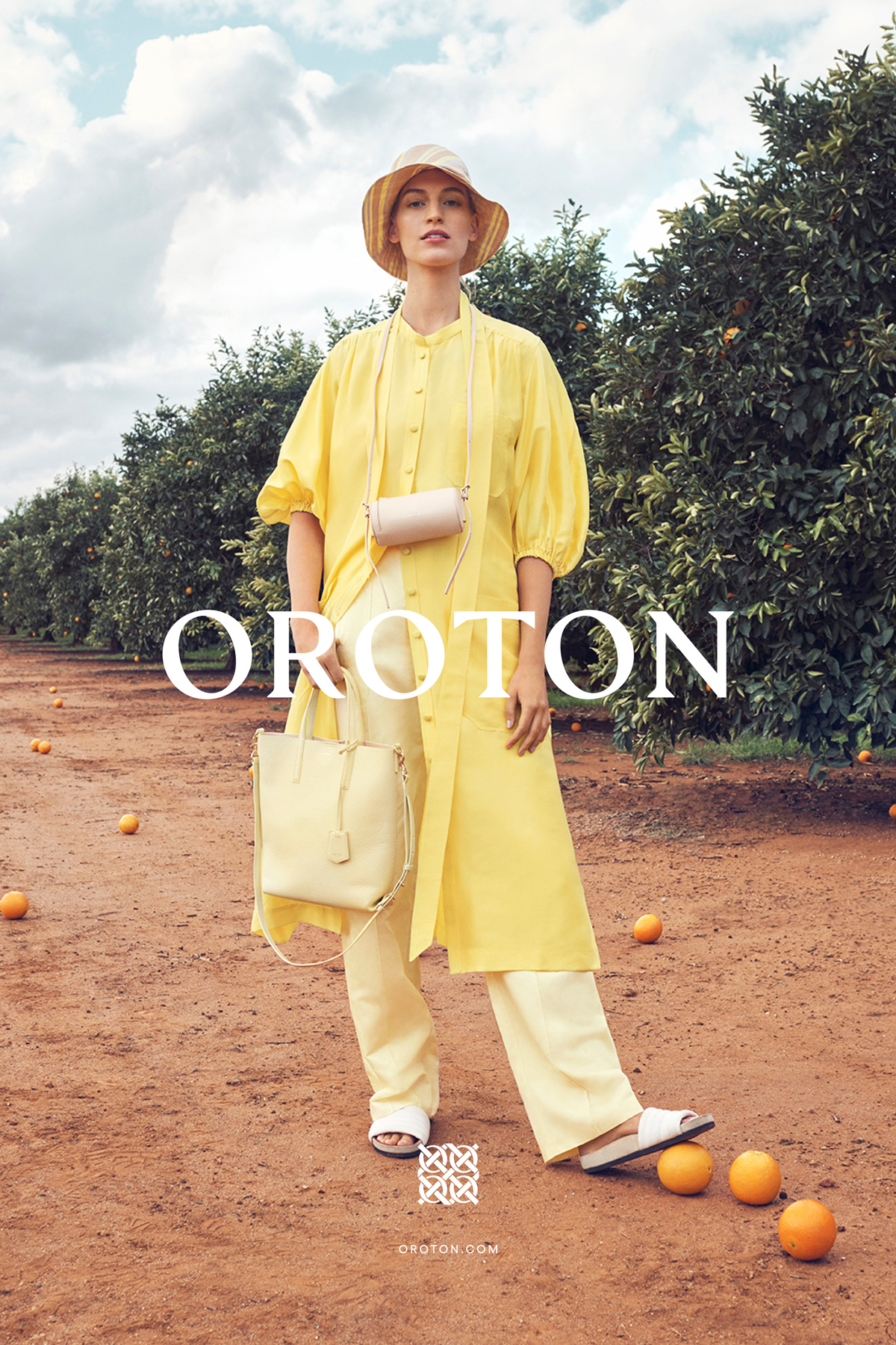 A blonde adult woman wears a yellow dress, hat, and white sandals while standing on a dirt path between rows of citrus trees.