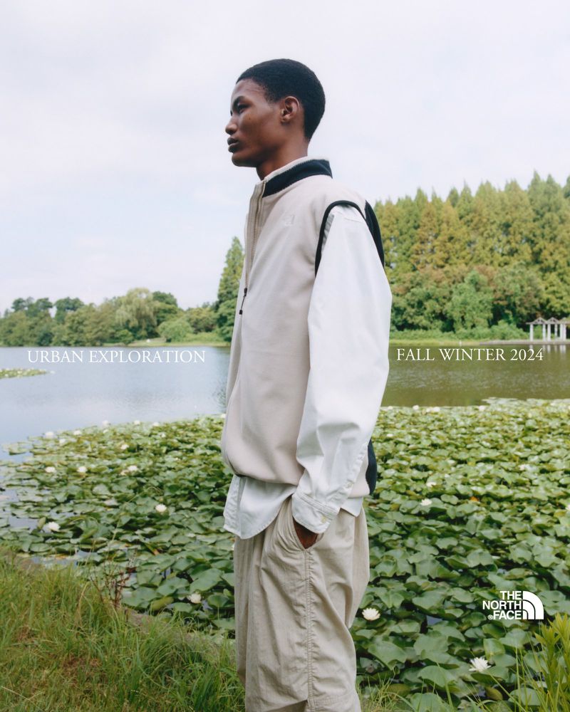 Outdoor fashion shoot featuring an adult Black male model in a neutral-toned jacket and trousers by a lake with lily pads, Fall Winter 2024.