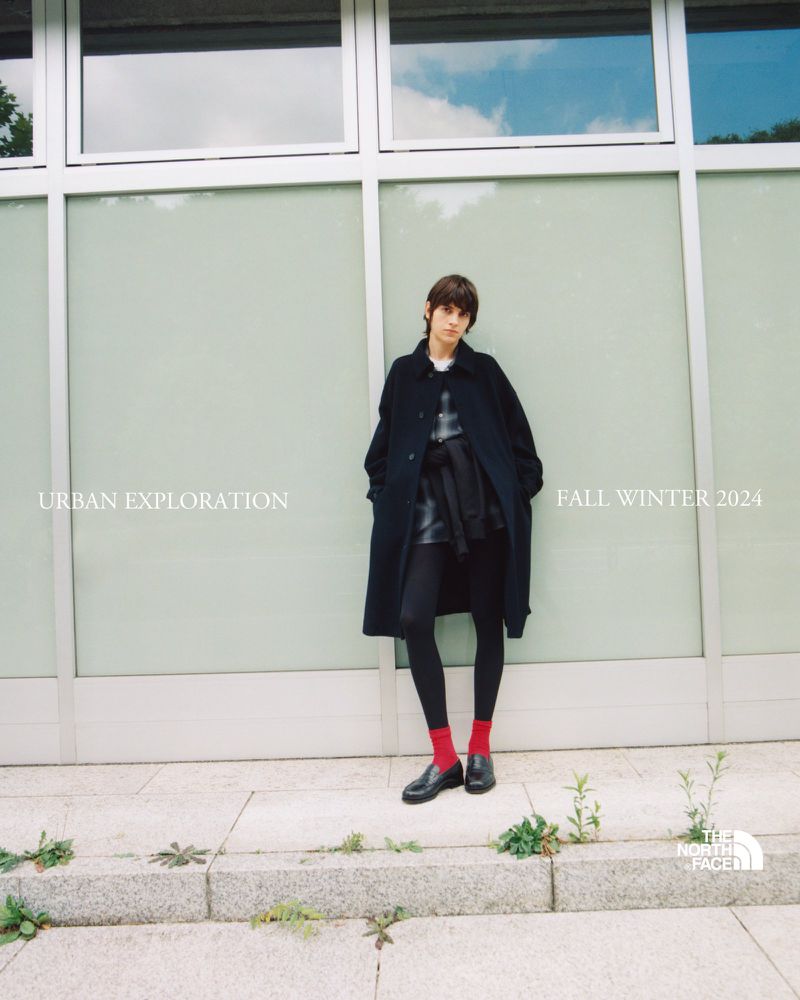 Fashion shot of a stylish young woman wearing a black coat, tights, and red socks, posing against a modern glass-walled building in an urban setting.