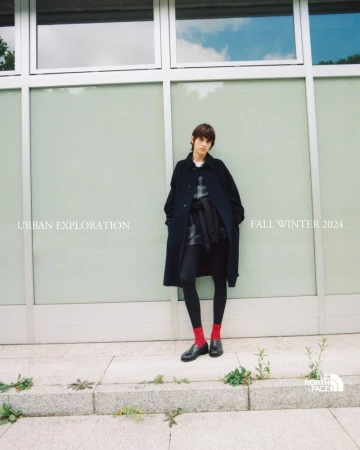 Fashion shot of a stylish young woman wearing a black coat, tights, and red socks, posing against a modern glass-walled building in an urban setting.
