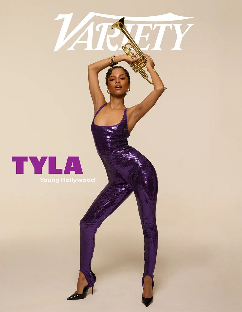 Full-length shot of Tyla, a Black woman in a sparkly purple jumpsuit, holding a trumpet overhead against a light-beige backdrop.