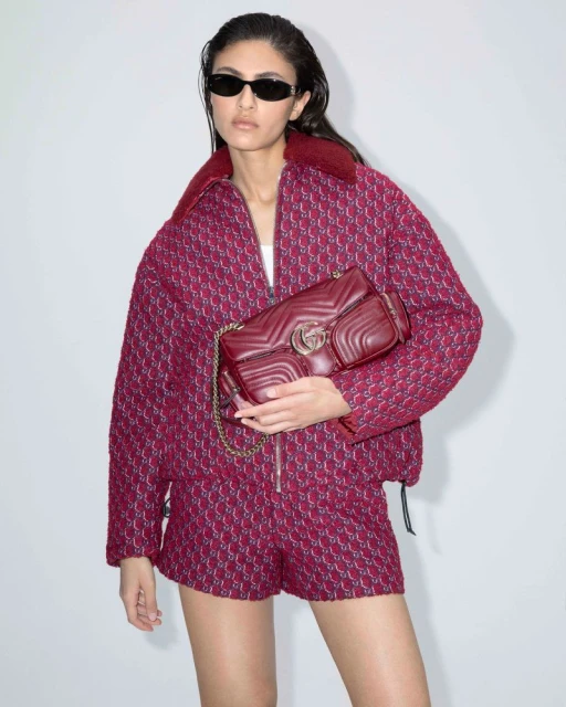 A fashionable adult female model poses in a patterned jacket and shorts with a leather purse against a white background in a studio.