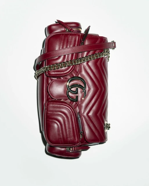 Studio shot of a burgundy leather Gucci Marmont backpack with chevron quilting, gold chain strap, and metal GG logo on a white background.