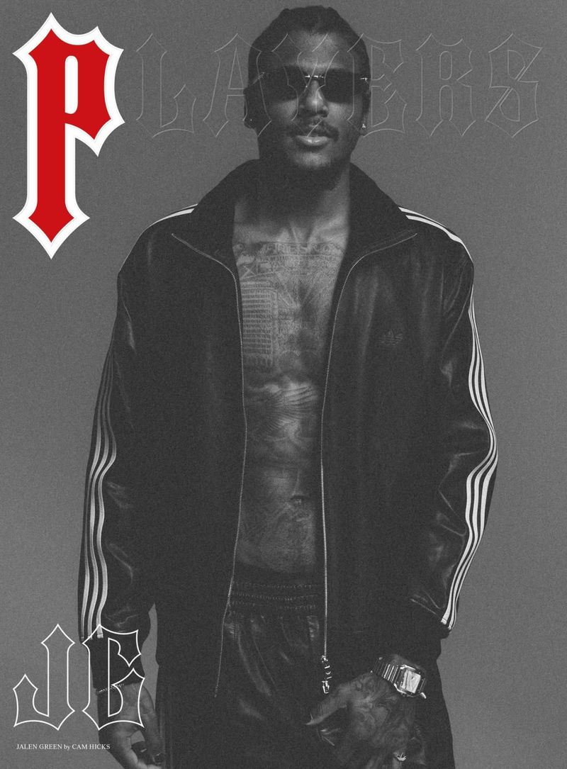 Monochrome portrait of a Black adult male with sunglasses, wearing a jacket, jewelry, and tattoos, against a neutral studio backdrop.