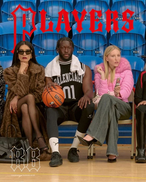 Three people sit on blue bleachers in a basketball arena. A man in a Balenciaga jersey holds a Wilson basketball.