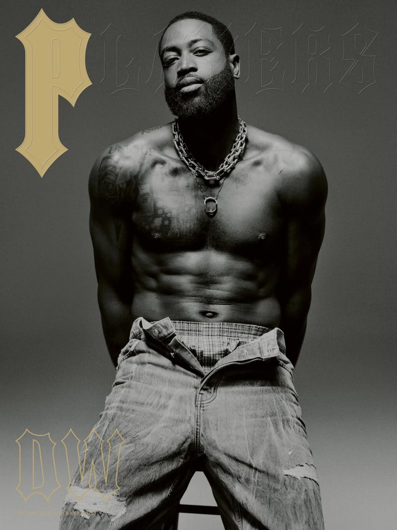 Monochrome portrait of a shirtless Black male model with beard, silver chain, and distressed jeans, posing on a stool in a studio.