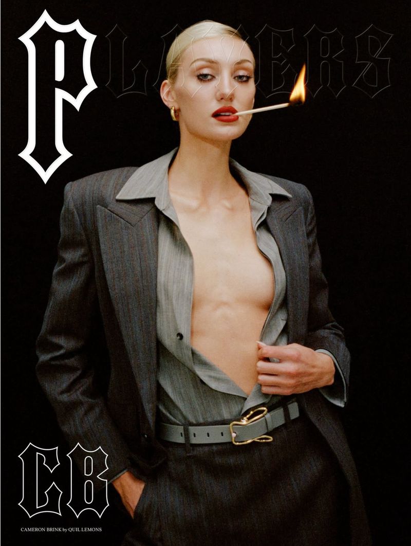Fashion editorial portrait of an adult female wearing a gray suit and holding a lit match in her mouth against a black background.