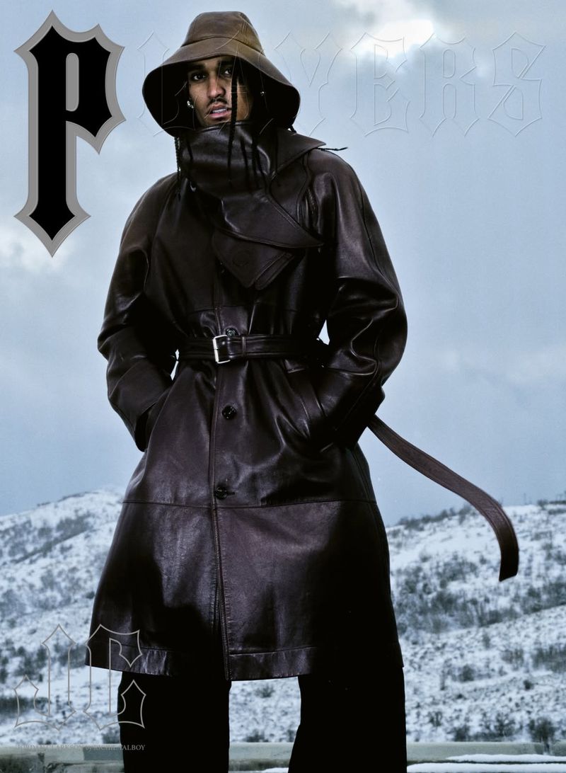 An adult male poses in a brown leather trench coat and bucket hat, against a background of mountains and overcast skies.