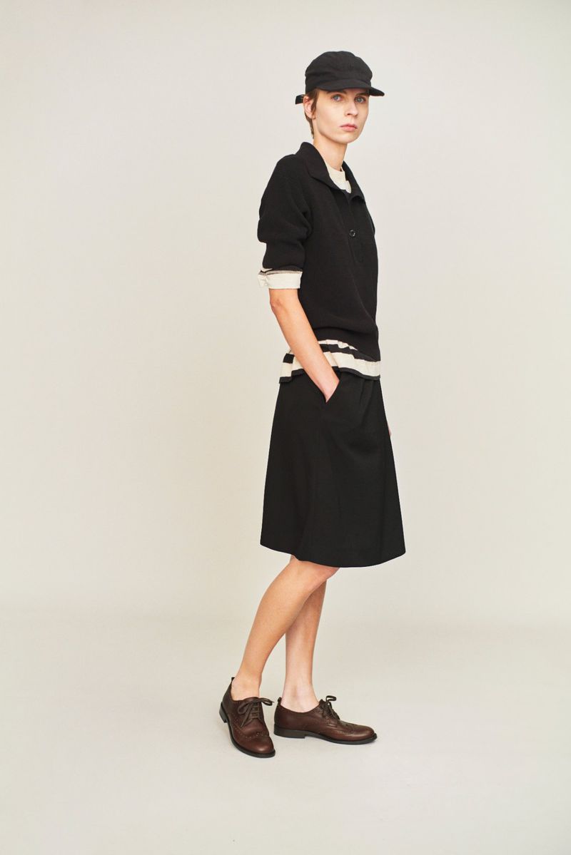 Full shot of an adult woman wearing a black shirt, skirt, and cap with brown dress shoes on a white background in a fashion setting.
