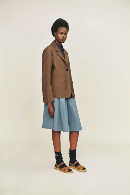 A brown-skinned model poses in a brown blazer, blue skirt, and brown sandals against a white background in a fashion setting.