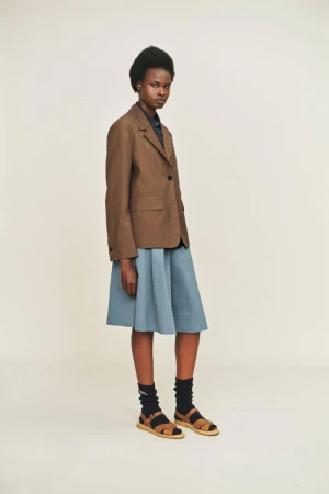 A brown-skinned model poses in a brown blazer, blue skirt, and brown sandals against a white background in a fashion setting.