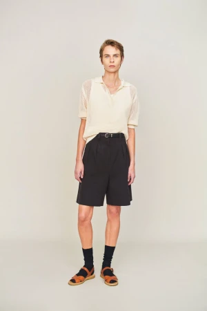 Model wearing a cream polo knit top and black shorts with dark socks and brown sandals against a neutral studio backdrop