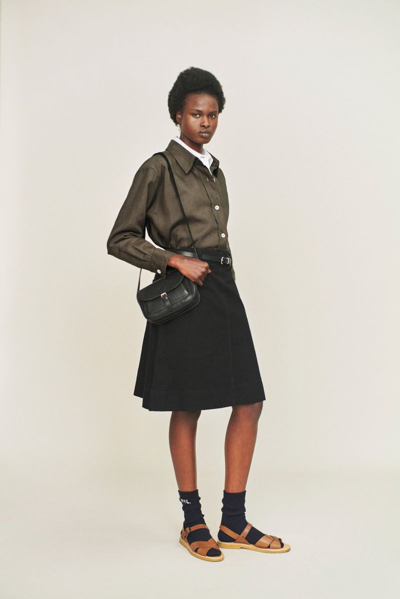 Model wearing an olive green shirt, black skirt, and leather crossbody bag standing against a white seamless backdrop.