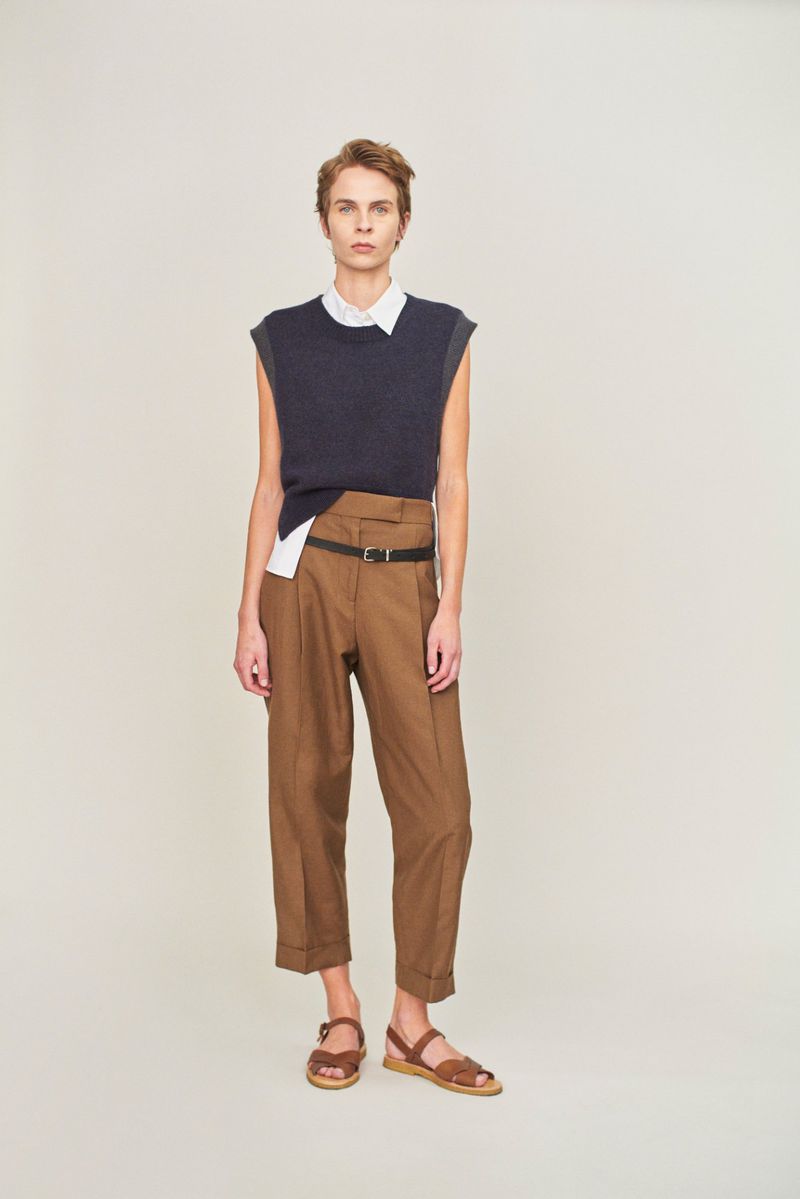 Full-length studio shot of an adult woman in a navy vest, white shirt, brown pants, brown belt, and brown sandals against white backdrop.