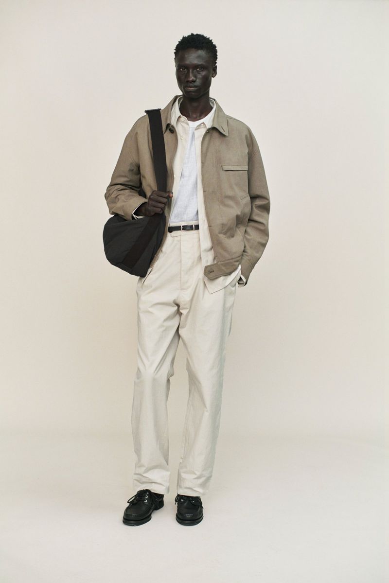 Model wearing a tan jacket, white shirt, and light trousers with a black crossbody bag against a neutral studio background