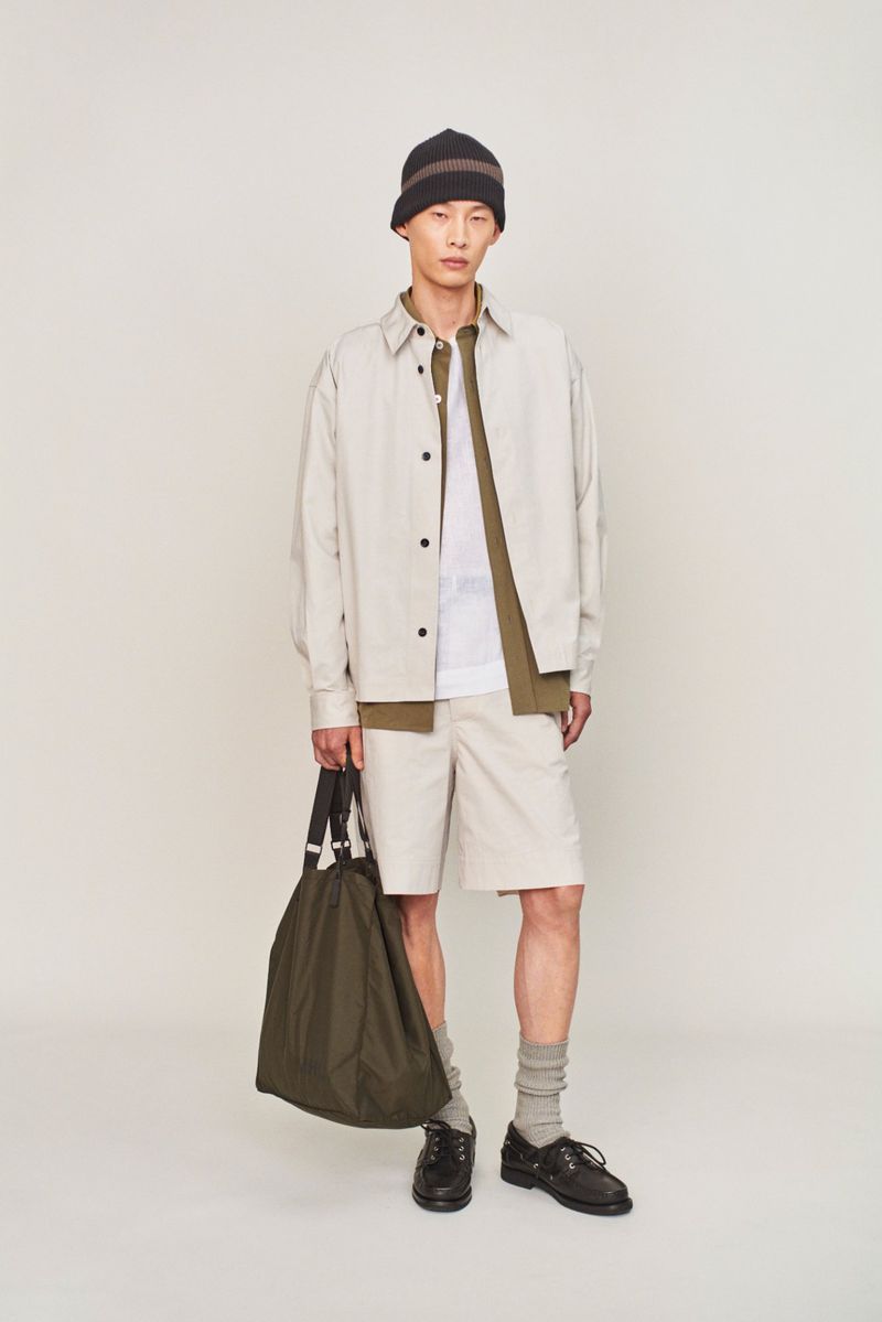 Young man in neutral-toned casual fashion, posing indoors against a white backdrop, holding olive-green tote bag, wearing a brown beanie.