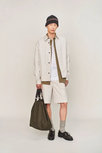 Young man in neutral-toned casual fashion, posing indoors against a white backdrop, holding olive-green tote bag, wearing a brown beanie.